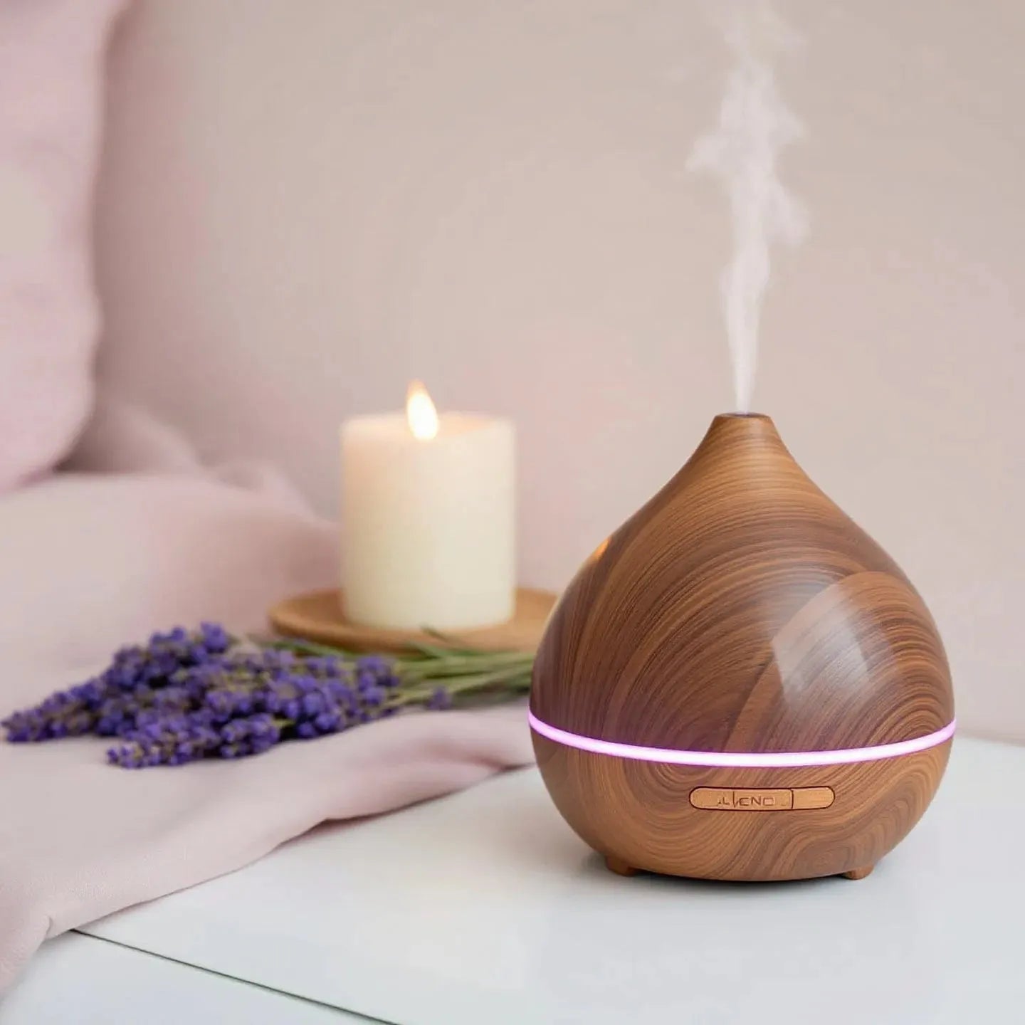 Ein stilvoller Holz-Diffuser, aus dem sanfter Dampf aufsteigt, steht auf einem weißen Tisch. Daneben liegen frische Lavendelblüten und eine brennende weiße Kerze auf einem Holzuntersetzer. Die Szene vermittelt eine beruhigende und entspannende Atmosphäre.