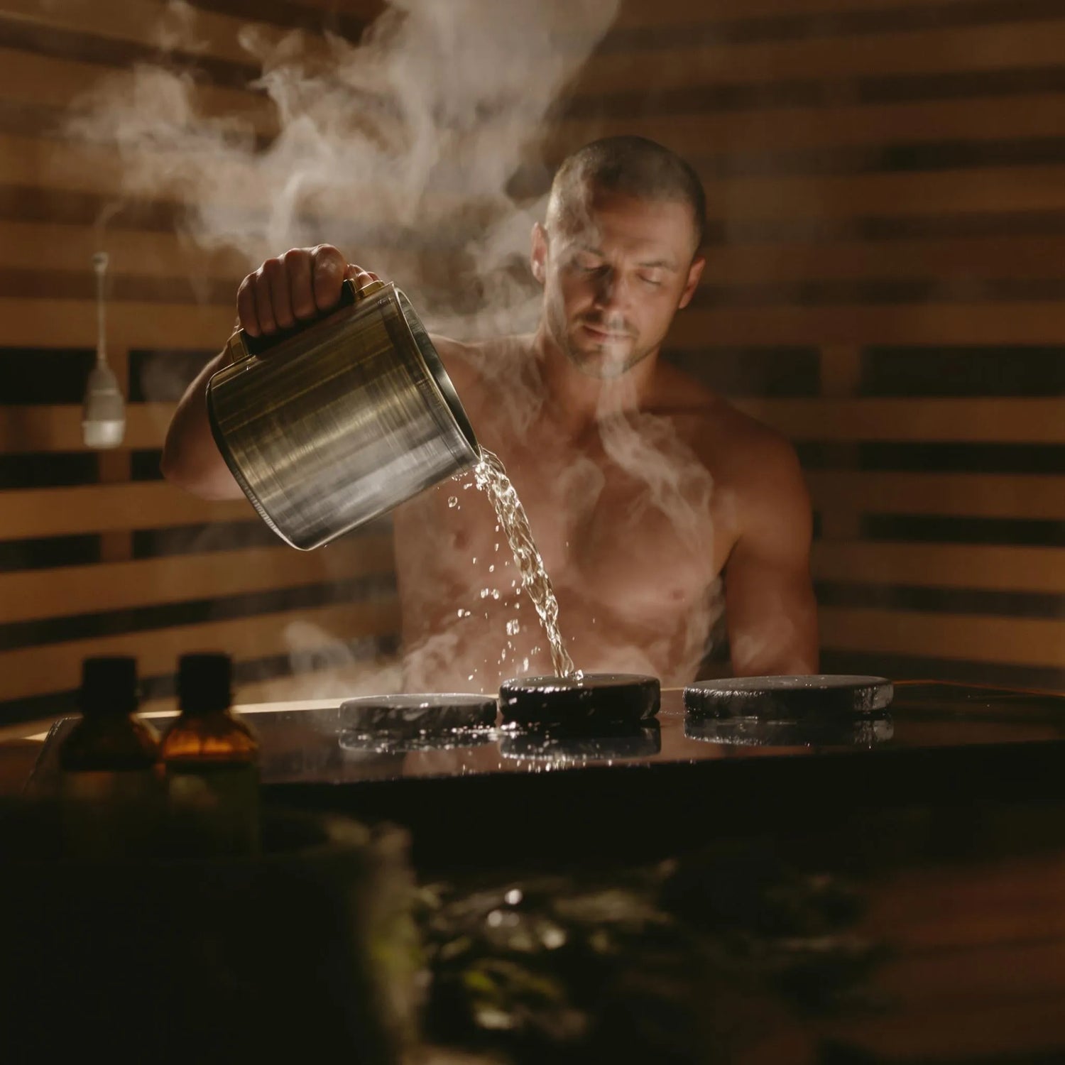 Mann gießt mit eine Kanne Wasser auf heiße Saunasteine für einen Saunaaufguss.