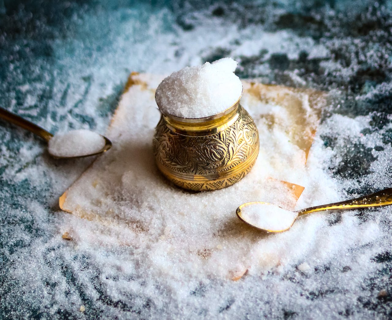 Mit Salz befülltes Kupferkännchen und zwei mit Salz befüllte Kupferlöffel auf einem Putzlappen, der auf einem mit Salz bedeckten Tisch liegt